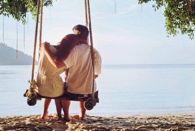 romantic holidays for two, affectionate couple sitting together on the beach on swing, silhouette of man hugging woman
